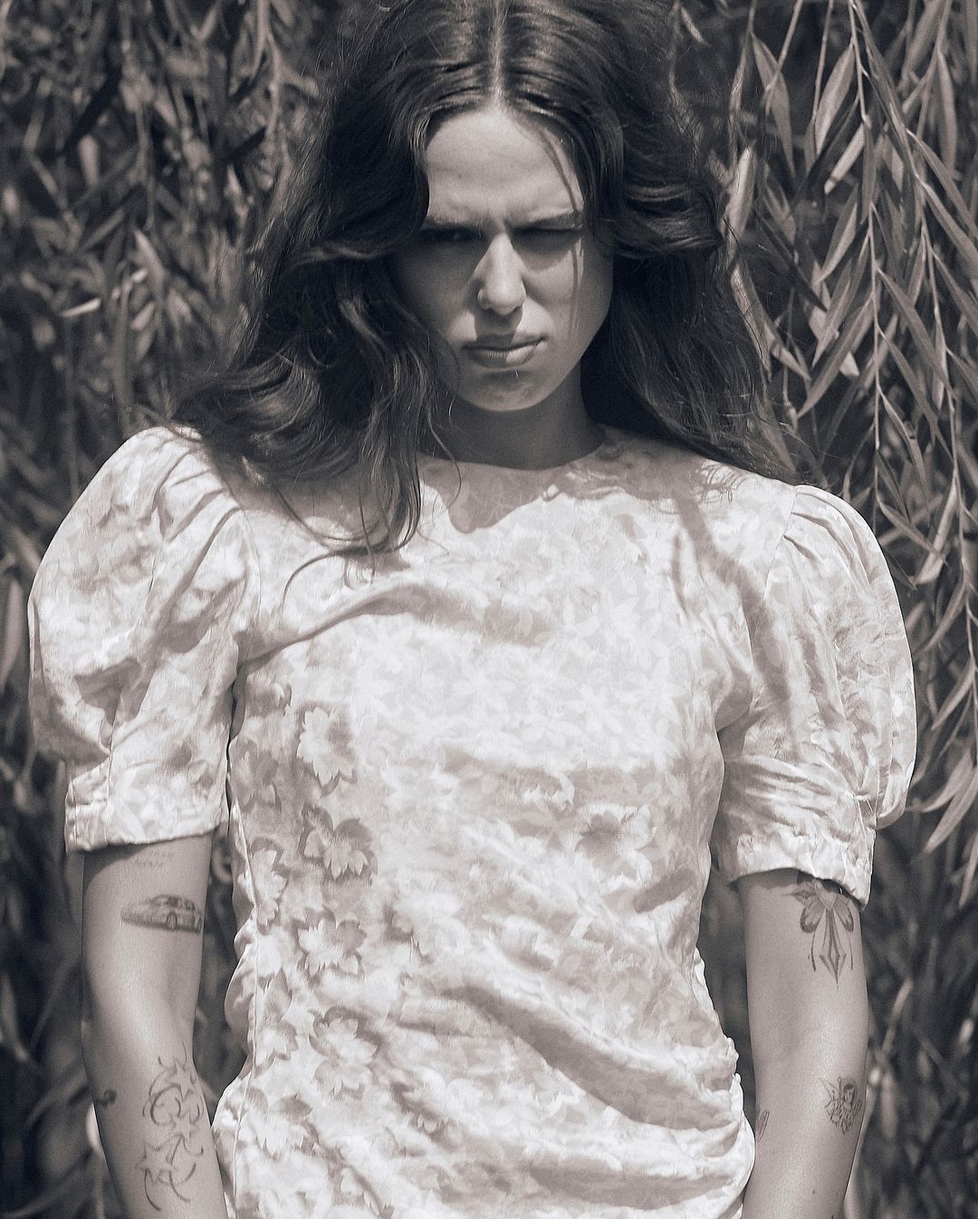 Monochrome portrait of a woman with long dark wavy hair wearing a patterned dress with puffed sleeves outdoors among foliage.