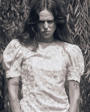 Monochrome portrait of a woman with long dark wavy hair wearing a patterned dress with puffed sleeves outdoors among foliage.