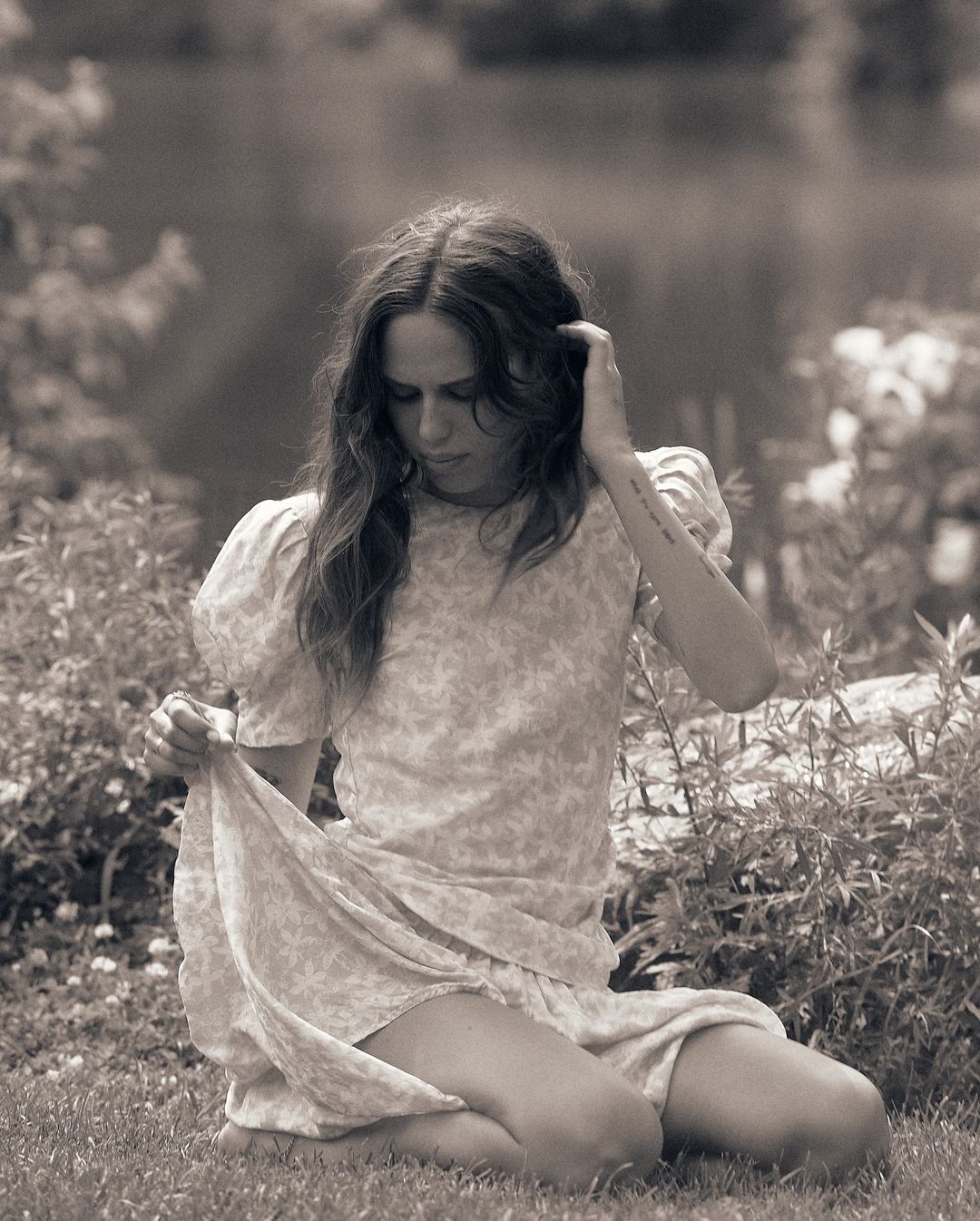 Woman in floral dress kneeling in a lush garden, adjusting the hem of her skirt while looking down outdoors