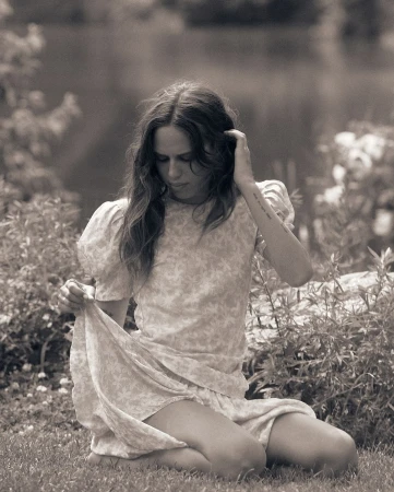 Woman in floral dress kneeling in a lush garden, adjusting the hem of her skirt while looking down outdoors