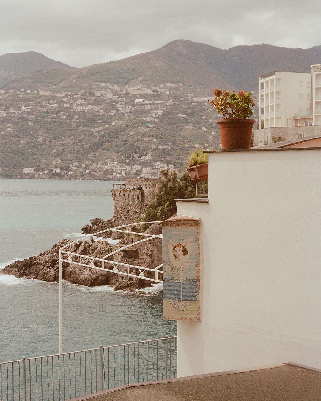 Seaside vista with a stone building on a rocky point, mountains in the background, and a potted plant atop a white building.