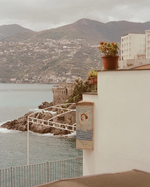 Seaside vista with a stone building on a rocky point, mountains in the background, and a potted plant atop a white building.