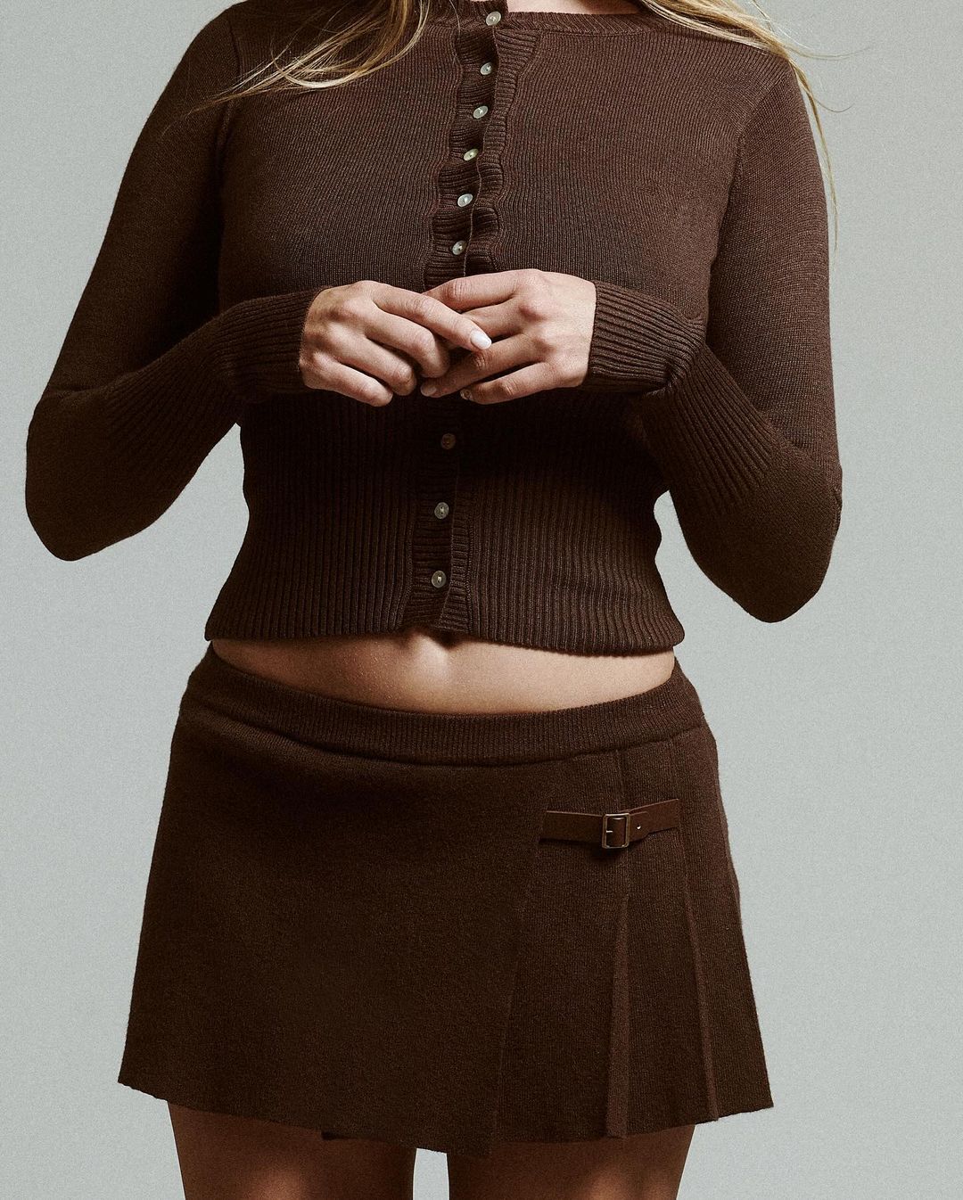 Woman wearing a brown knitted cardigan and a matching mini skirt in a studio setting with a soft grey background, fashion shot.