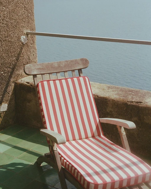Outdoor patio with a wooden lounge chair featuring a red and white striped cushion, overlooking the sea and a stone wall.