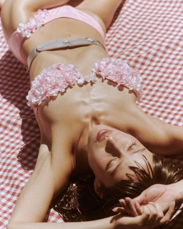Woman in pink floral bikini lying on a red-and-white checkered blanket outdoors, eyes closed, relaxed pose, sunny day, fashion shot.