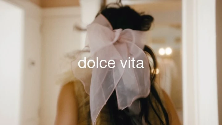 Woman with a pink bow in her hair, indoors with soft lighting. The focus is on fashion with the text “dolce vita” overlaid.
