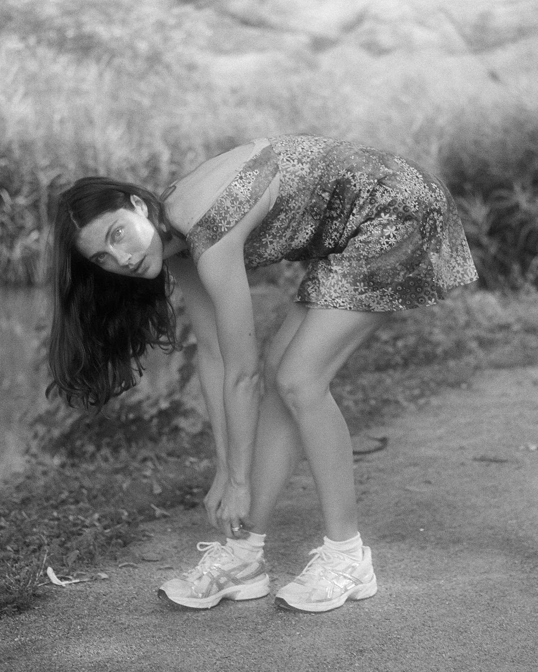 Black and white photo of a young woman tying her sneaker laces in a park setting, wearing a floral dress and white running shoes.