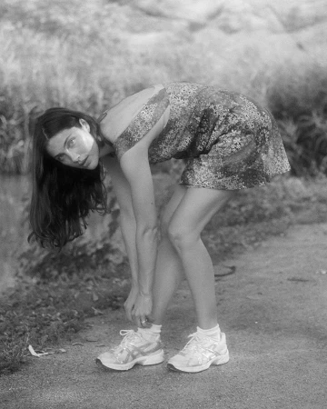 Black and white photo of a young woman tying her sneaker laces in a park setting, wearing a floral dress and white running shoes.