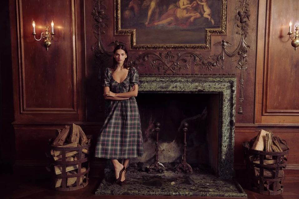 A woman in a plaid dress stands in front of a marble fireplace with wood baskets, vintage lighting, and a framed painting.
