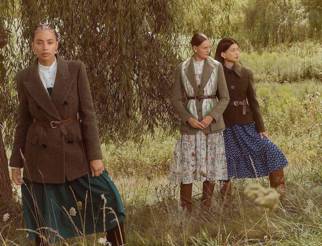 Three women wearing blazers and dresses pose outdoors in a grassy field with trees. Fashionable clothing, elegant style.