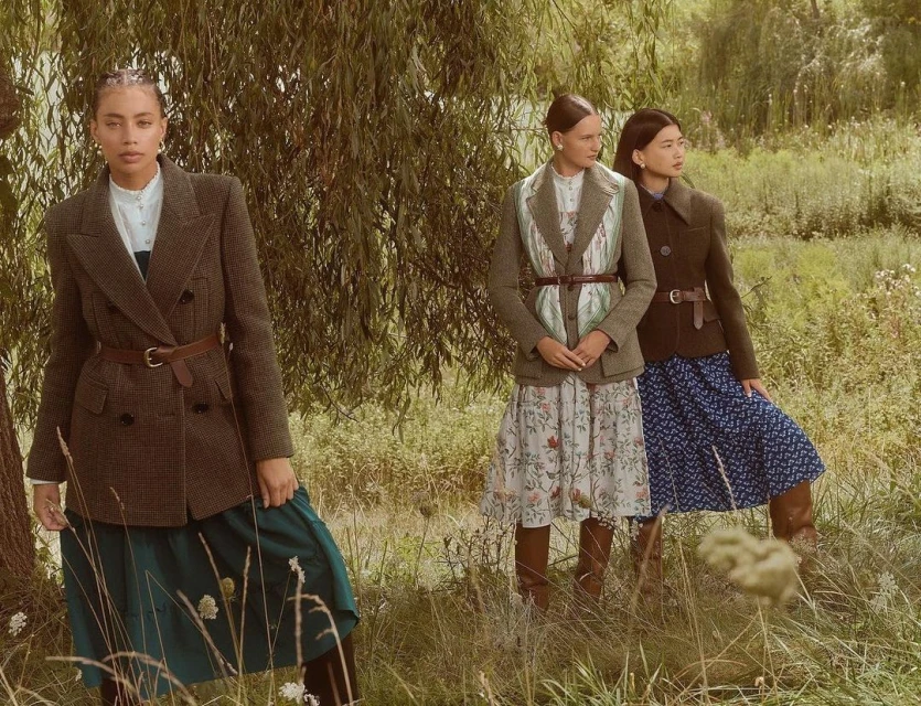 Three women wearing blazers and dresses pose outdoors in a grassy field with trees. Fashionable clothing, elegant style.