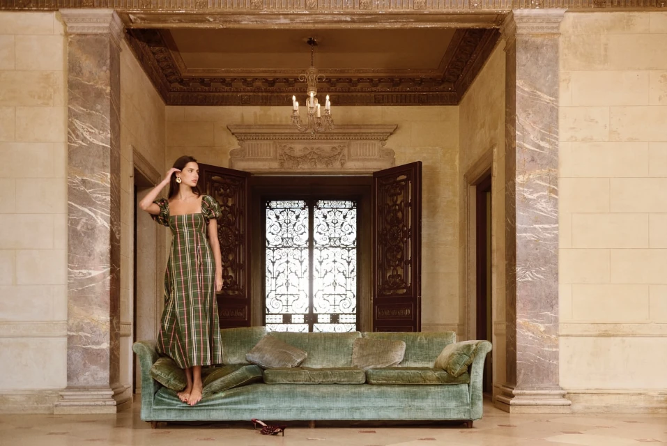 A barefoot adult woman in a green plaid dress posing on a green velvet couch with marble columns in a classically designed room.