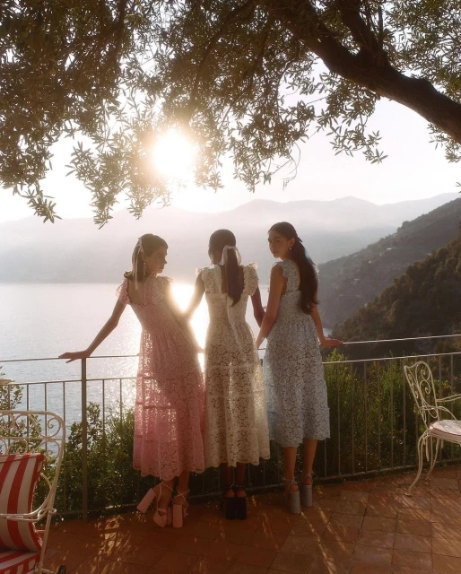 Three adult females in dresses standing on a terrace overlooking a sea view with mountains in the background, bathed in golden sunlight.