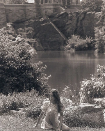 Woman with long hair wearing a patterned dress seated outdoors on a grassy lawn near a park pond