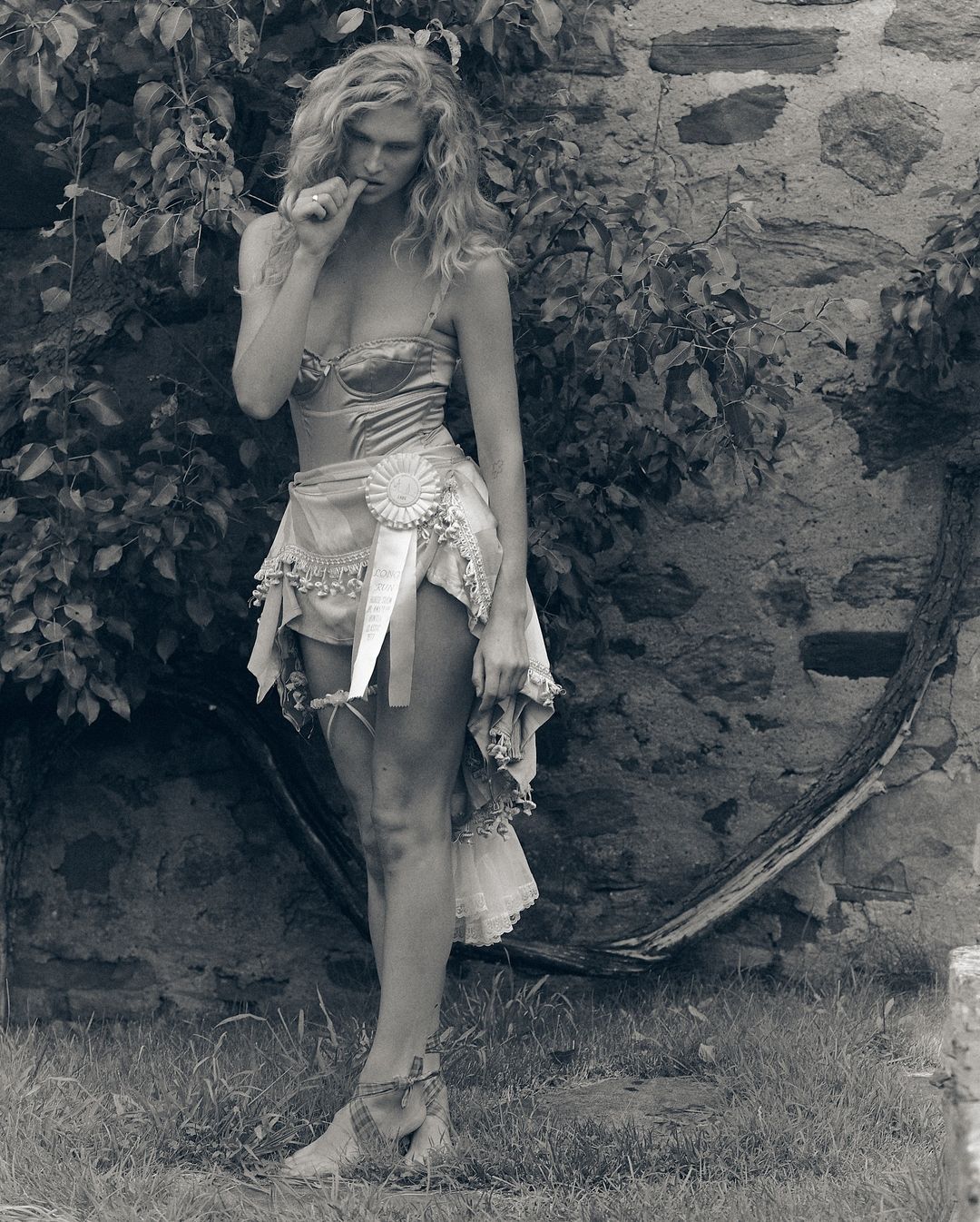 Model with long blonde hair wearing a corset and draped skirt posing outdoors against a rustic stone wall and tree branches