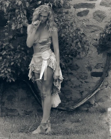 Model with long blonde hair wearing a corset and draped skirt posing outdoors against a rustic stone wall and tree branches