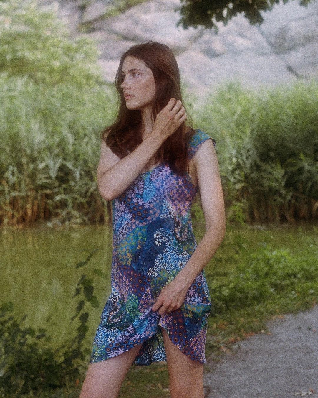 Young adult woman in a floral sundress stands in a park landscape with a pond and trees in soft, diffused natural light.