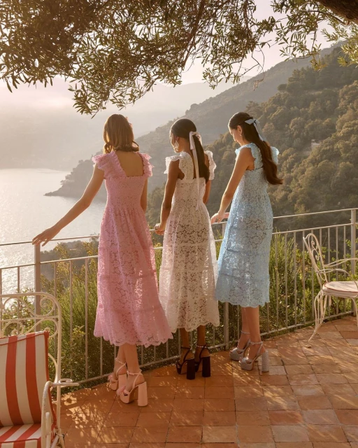 Three women in pastel lace dresses stand on a balcony overlooking a scenic vista with mountains and water during daytime.
