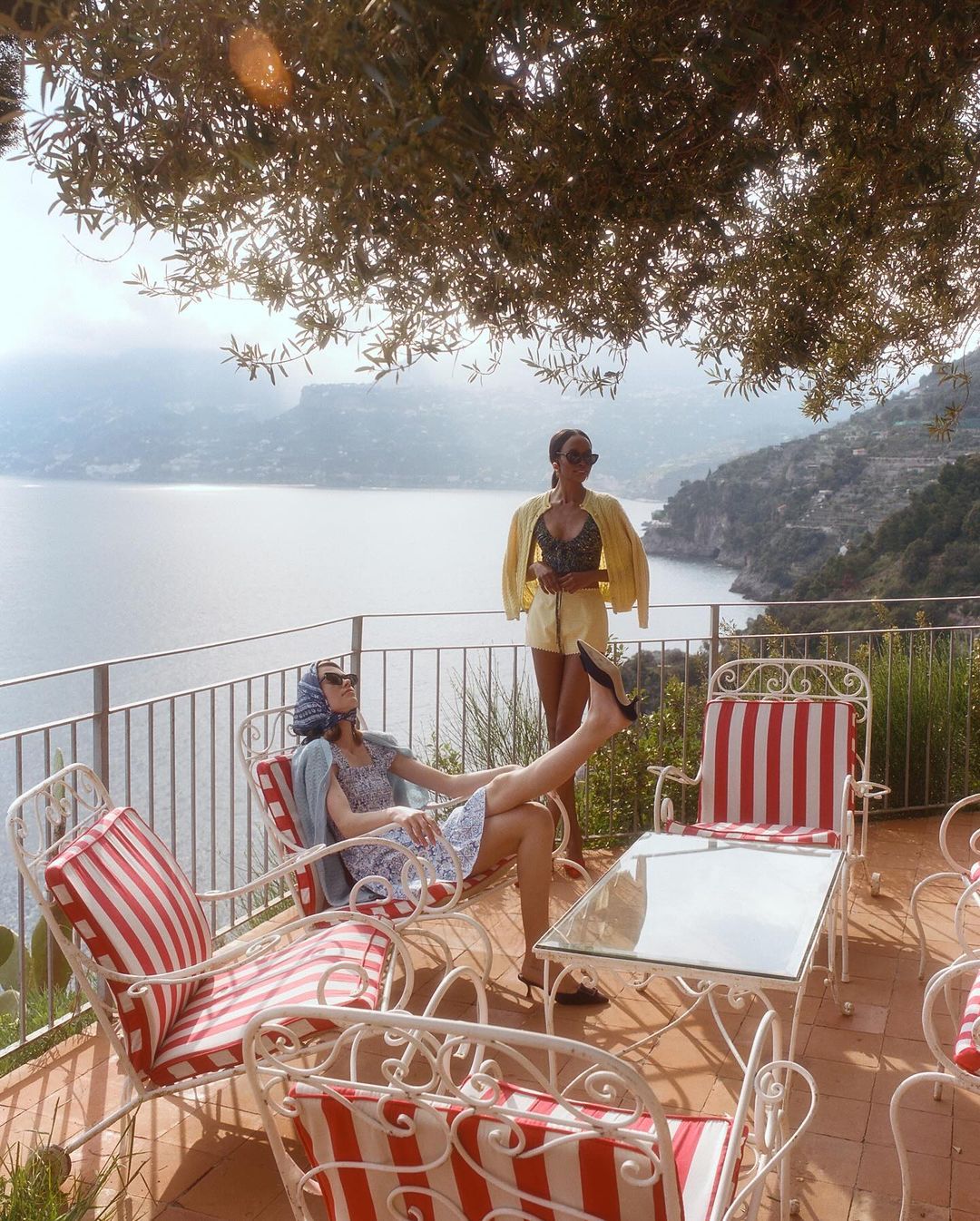 Two women on a terrace overlooking the sea. One is reclining in a striped chair and the other is standing with a yellow jacket.