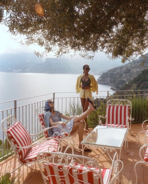 Two women on a terrace overlooking the sea. One is reclining in a striped chair and the other is standing with a yellow jacket.