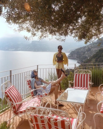 Two women on a terrace overlooking the sea. One is reclining in a striped chair and the other is standing with a yellow jacket.