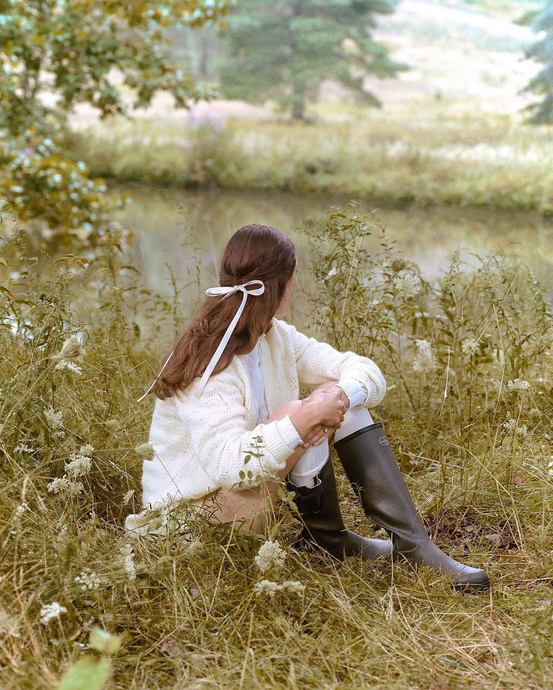 A young woman with a white bow in her hair sits near a grassy riverbank on a sunny day, wearing a cream sweater and tall boots.