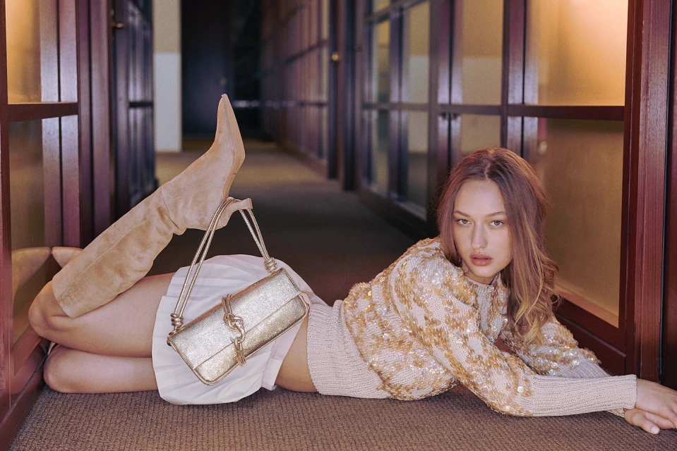 Woman lying on the floor with a purse and suede boots wearing a sequin sweater and skirt in a hallway with wood paneled doors.