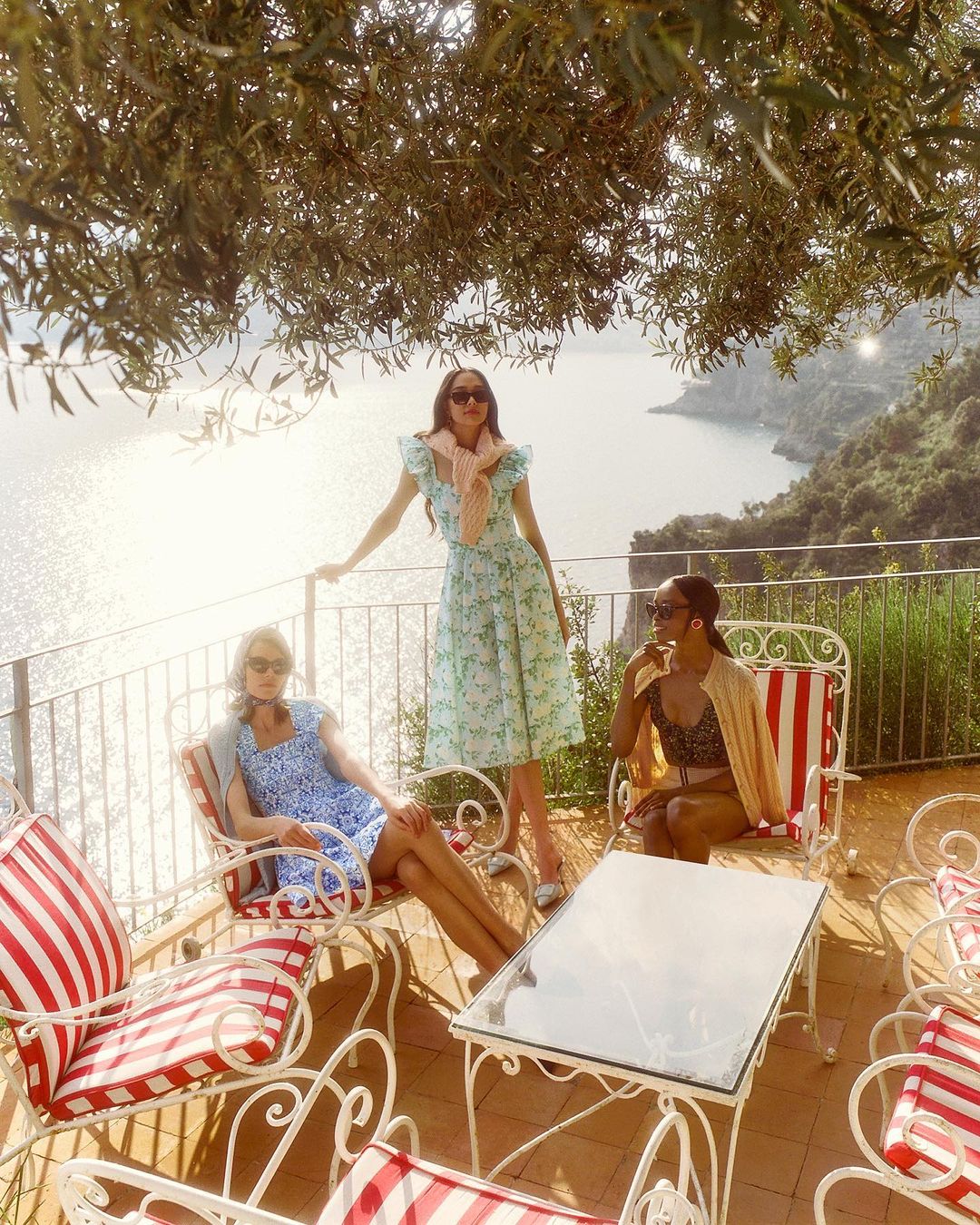 Three women pose on a sunlit terrace overlooking the sea. One stands in a floral dress, while two others relax in red-striped chairs.