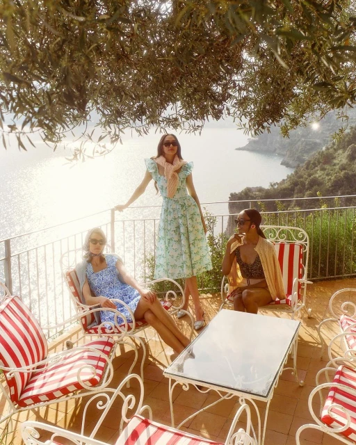 Three women pose on a sunlit terrace overlooking the sea. One stands in a floral dress, while two others relax in red-striped chairs.