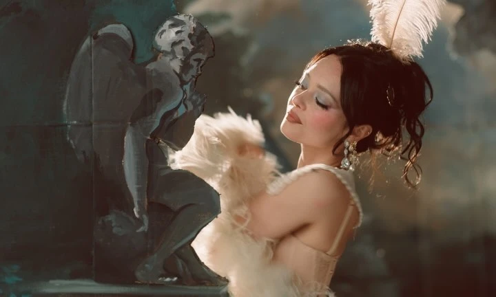 Glamorous woman with feather accessory posing beside a painting of a seated angel in an indoor studio setting with warm lighting.