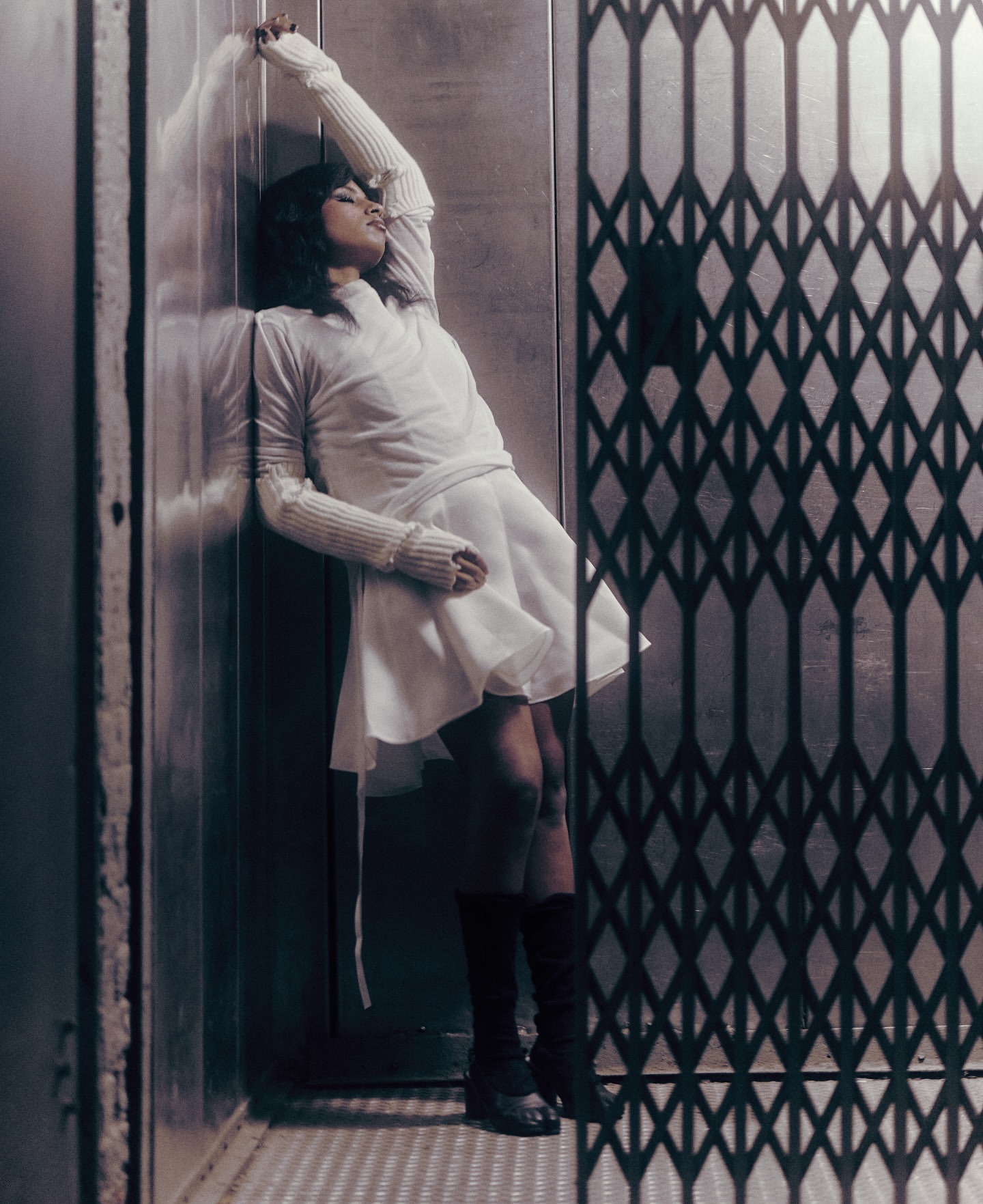 Stylish woman in a white dress poses dramatically in an elevator with a wrought-iron gate, creating a vintage fashion statement.
