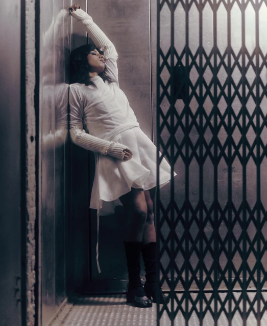 Stylish woman in a white dress poses dramatically in an elevator with a wrought-iron gate, creating a vintage fashion statement.