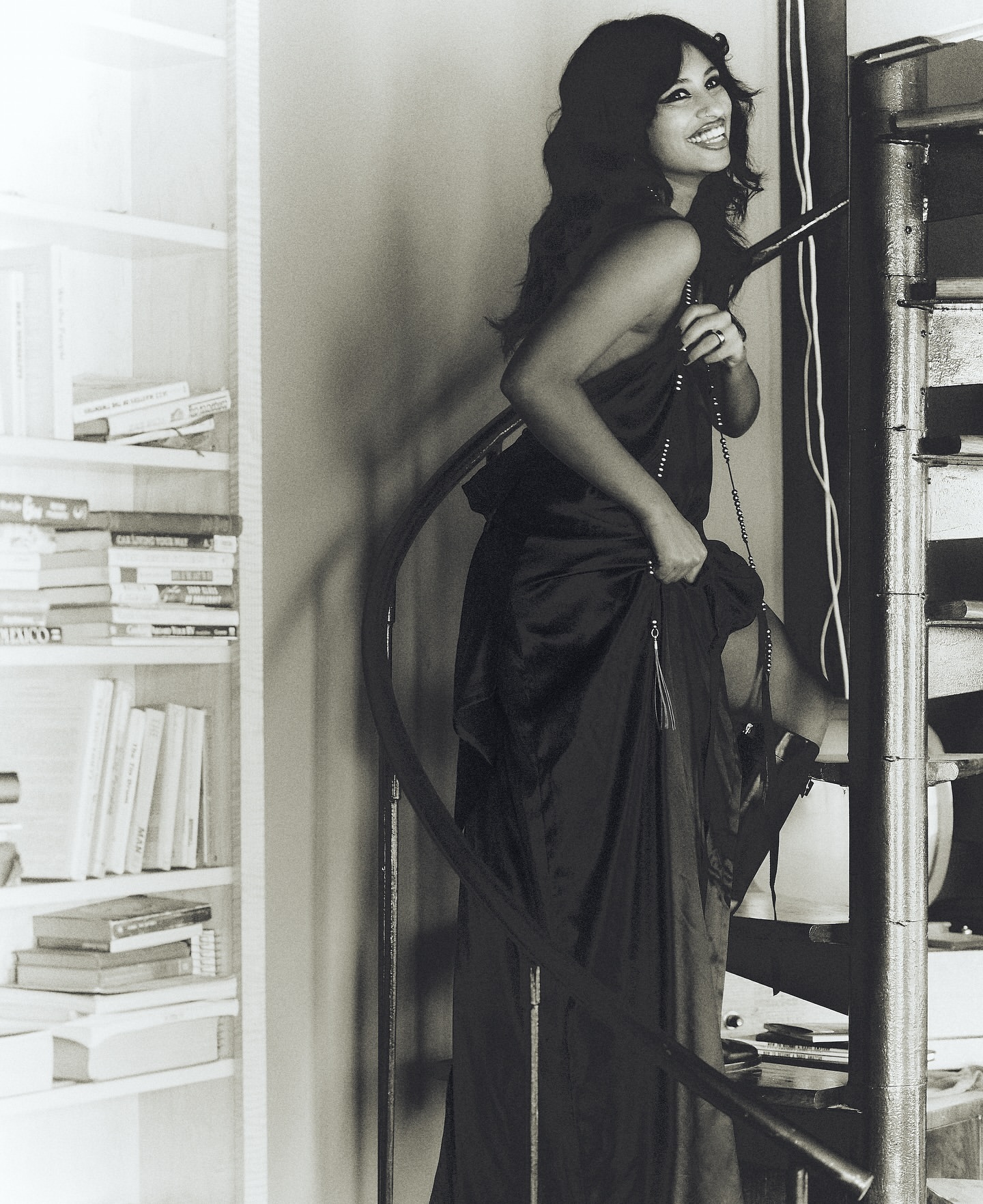 A woman in a long black dress stands on a winding staircase next to a white bookshelf, smiling and looking at the camera.