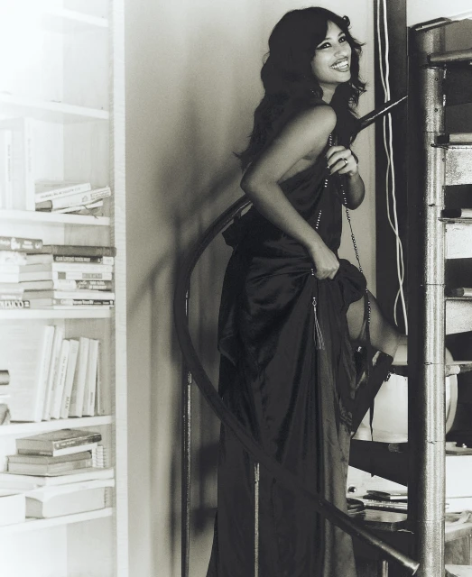 A woman in a long black dress stands on a winding staircase next to a white bookshelf, smiling and looking at the camera.