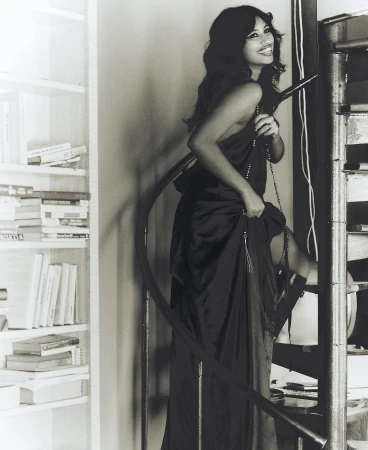A woman in a long black dress stands on a winding staircase next to a white bookshelf, smiling and looking at the camera.