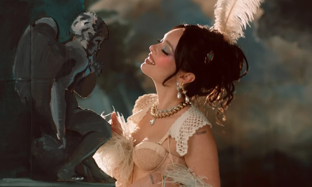 Adult female posing in profile with artistic statue backdrop, wearing period-style clothing and feather hair accessory, in eye-level shot.
