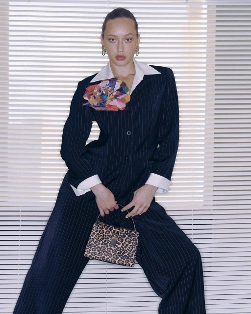 A woman poses in a pinstripe suit with a floral accent and leopard print handbag against a white slat wall, exuding fashion and style.