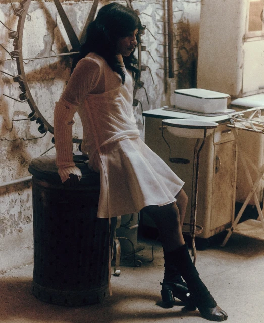Woman in a light-colored dress and black leg warmers sits on a metal trash can in an indoor setting with distressed walls and metal furniture.
