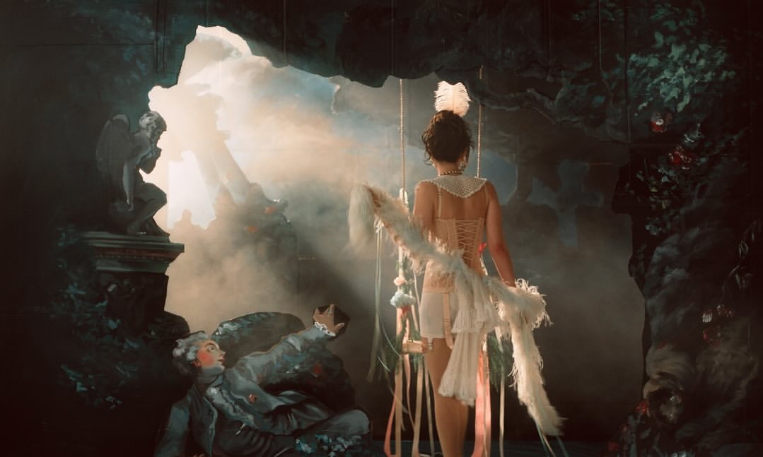 A woman wearing white lingerie and a feather boa stands on a stage with statues, theatrical lighting, and a surreal background.