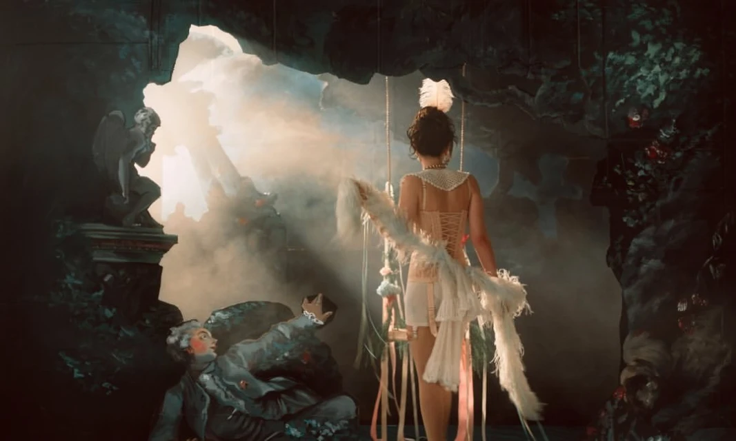 A woman wearing white lingerie and a feather boa stands on a stage with statues, theatrical lighting, and a surreal background.