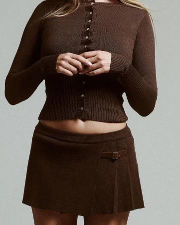Woman wearing a brown buttoned cardigan and matching miniskirt against a plain grey textured studio background.