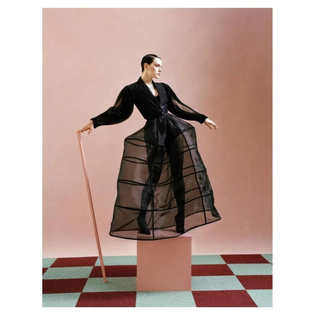 Fashionable adult female model in a black semi-sheer outfit poses on a block in a studio with a checkered floor and pink backdrop.