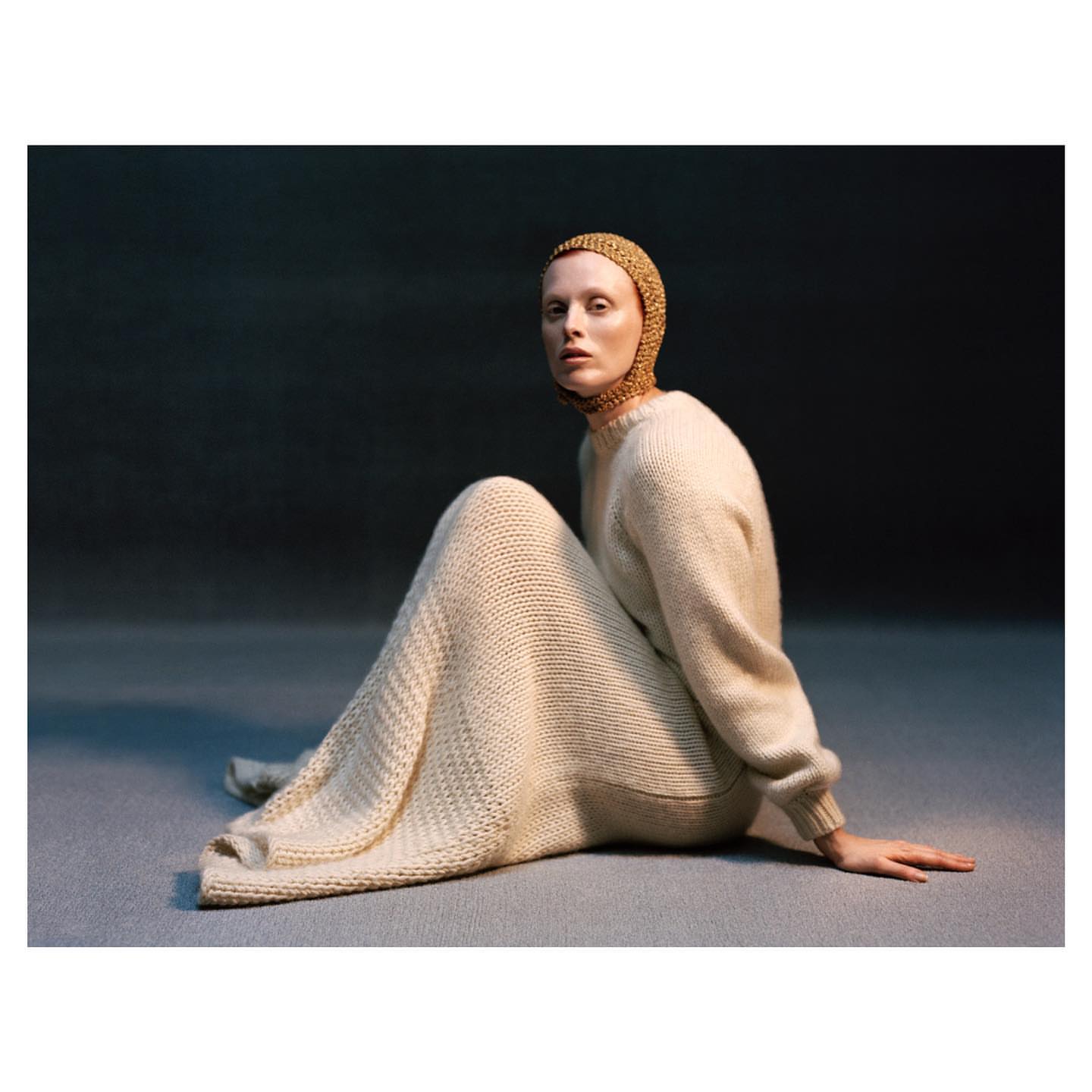 A pale-skinned woman sits on a gray surface, wearing a knitted cap and a long, knitted dress in a studio setting against a dark background.