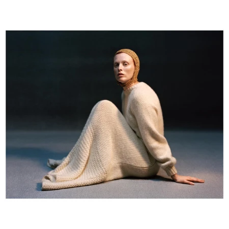 A pale-skinned woman sits on a gray surface, wearing a knitted cap and a long, knitted dress in a studio setting against a dark background.