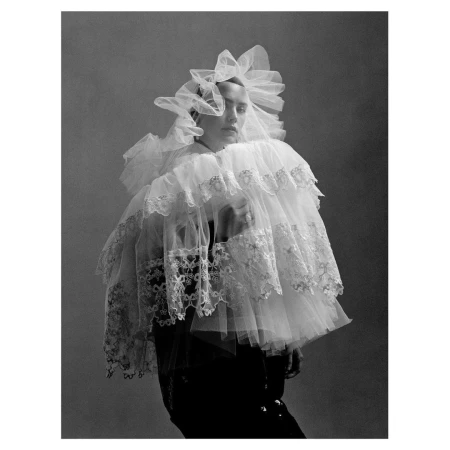 Monochrome portrait of a woman in a large, frilly white lace and tulle collar and headdress against a muted grey backdrop in studio lighting.