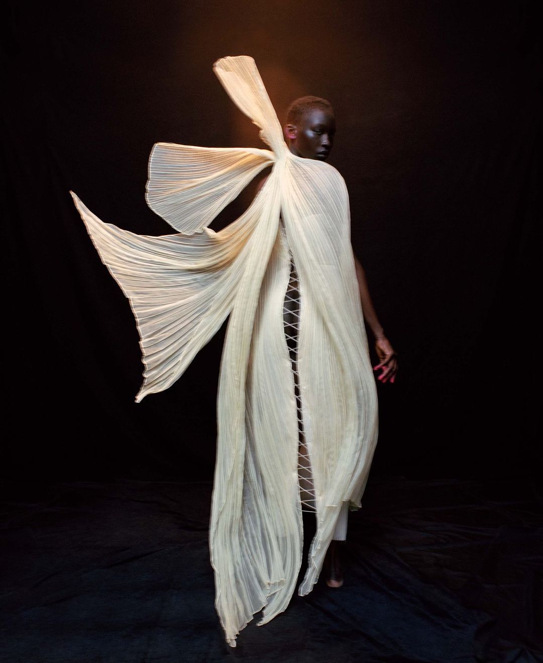 A model wears a flowing cream-colored dress with a dramatic pleated design against a dark background in a fashion-forward pose.