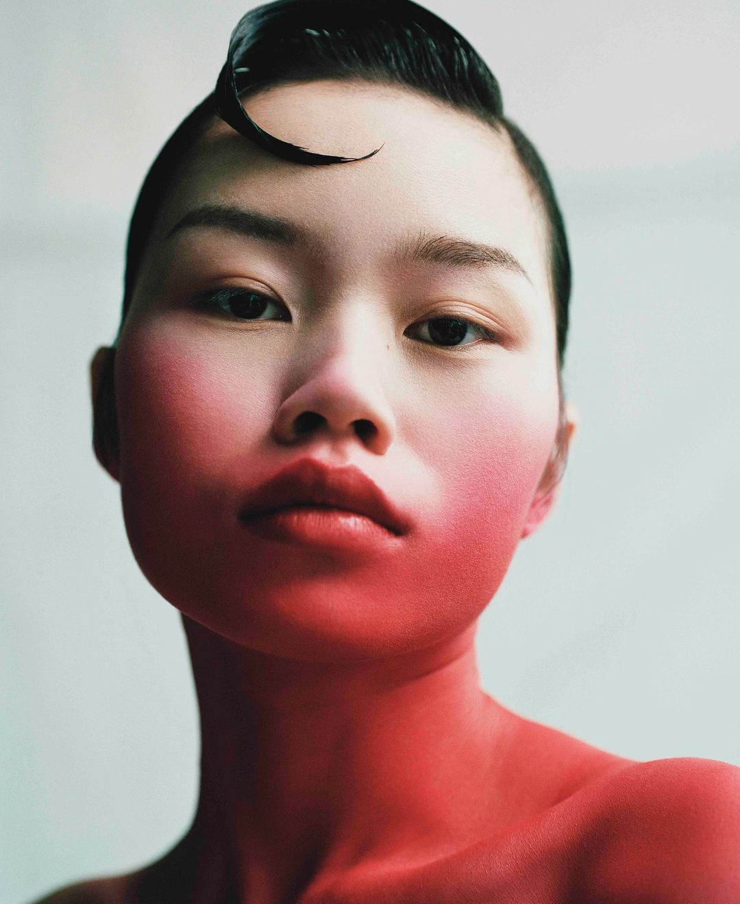 Close-up portrait of an Asian woman with styled hair, wearing minimal makeup, and posing against a plain background with soft, diffused lighting.