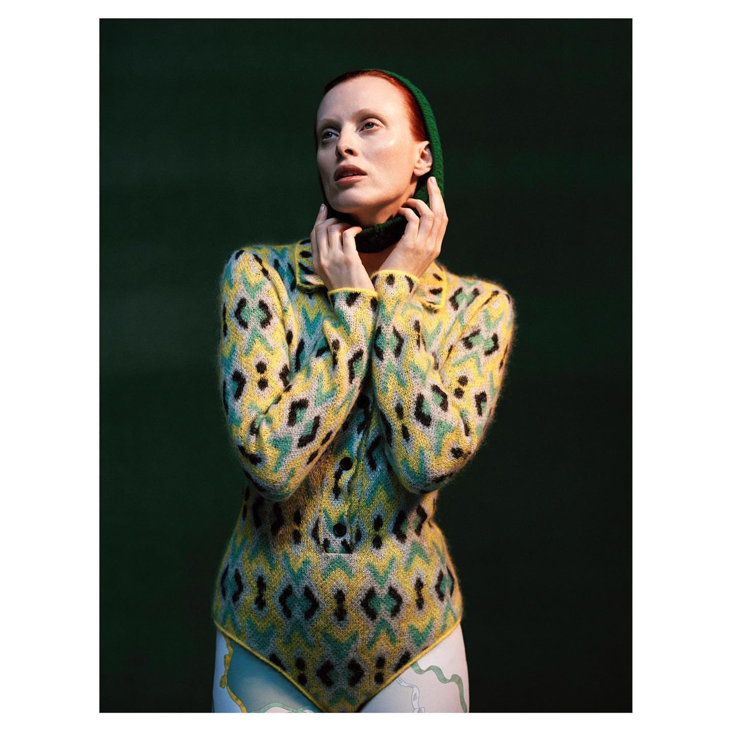 A fair-skinned adult female with red hair poses in a patterned yellow and black sweater and white bottoms against a dark green backdrop.