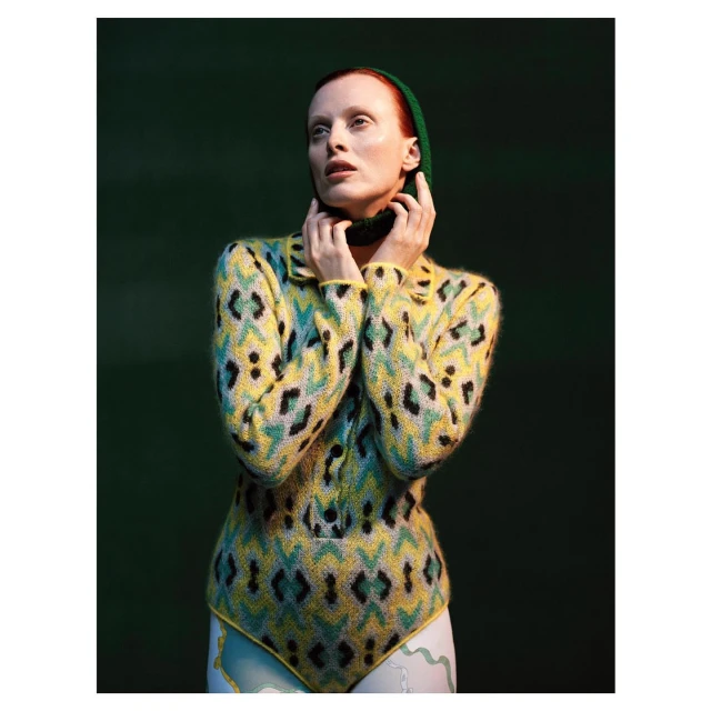 A fair-skinned adult female with red hair poses in a patterned yellow and black sweater and white bottoms against a dark green backdrop.