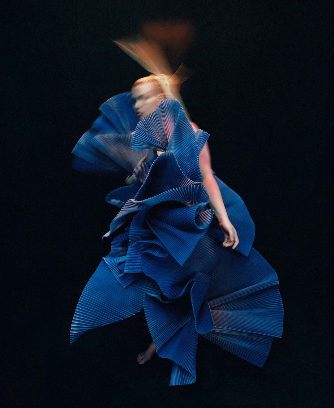 Woman wearing an elaborate, pleated blue dress in motion against a dark background, captured with intentional motion blur for artistic effect.